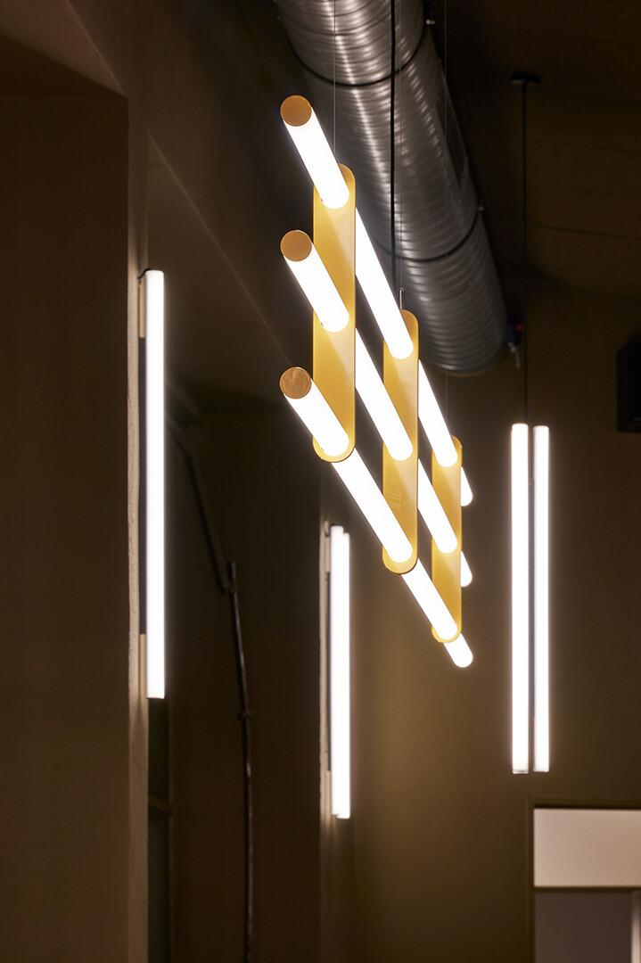 Moderne, minimalistische Lobby-Beleuchtung mit einer skulpturalen Pendelleuchte aus gelben Metallelementen und LED-Lichtröhren in einem industriell gestalteten Empfangsbereich.